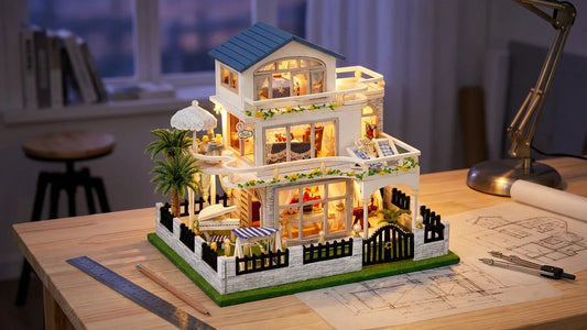 La Villa de la Riviera montée et allumée sur une table d'architecte claire, à côté d'une règle en métal, d'un crayon, d'un compas et de plans de maison dessinés au crayon, sous une lampe de bureau, les trois niveaux éclairés pièce par pièce