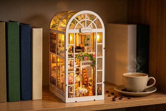 Le book nook Le Jardin de Café allumé sur une étagère en bois, entre des livres aux dos unis, avec sa verrière en arche et ses enseignes, à côté d'une tasse de café fumante et de grains de café