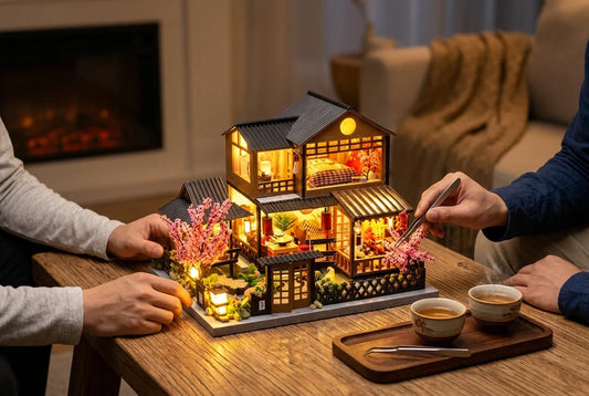 La maison miniature japonaise La Maison aux Cerisiers allumée sur une table en bois, le soir : deux paires de mains en manches longues la terminent ensemble, l'une pose une pièce avec une pince, à côté de deux tasses de thé