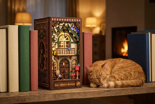 Le book nook Le Bureau des Affaires Félines allumé entre des livres aux dos unis sur une étagère en bois, le soir, avec un vrai chat roux endormi en boule juste à côté et un feu de cheminée au fond