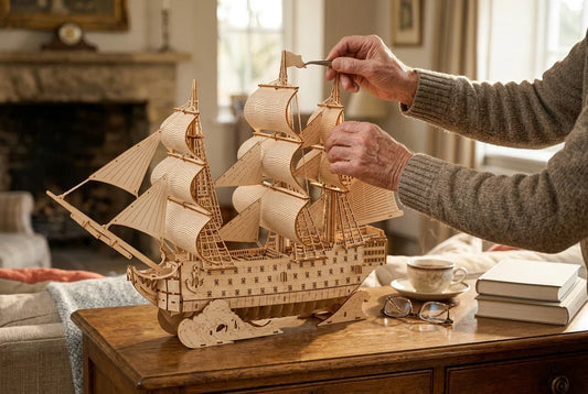 La maquette en bois Le Vaisseau Amiral posée sur un buffet ancien dans un salon chaleureux : deux mains en pull de laine ajustent un drapeau du mât avec une pince fine, à côté de lunettes et d'une tasse de thé