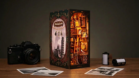 Le book nook La Chambre Noire allumé sur un bureau en bois le soir, entre un appareil photo argentique noir, deux rouleaux de pellicule et des tirages noir et blanc posés à plat