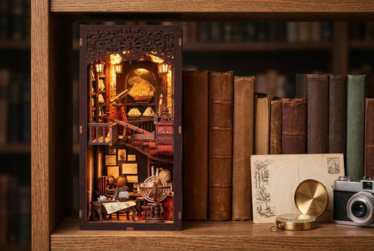 Le book nook Le Carnet de Voyage monté et allumé sur une étagère, entre des livres anciens, à côté d'une carte postale, d'une boussole en laiton et d'un appareil photo argentique