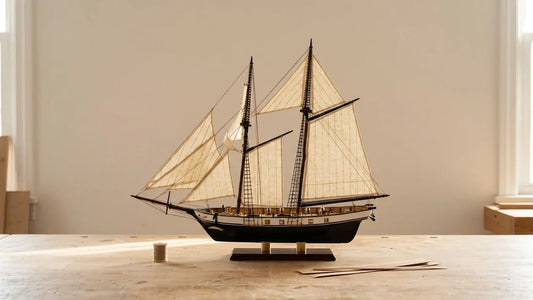 La maquette du Voilier Harvey terminée sur un établi en bois clair dans un atelier en fin de journée : goélette à deux mâts, coque noire et blanche, voiles en toile, bobine de fil et baguettes à côté