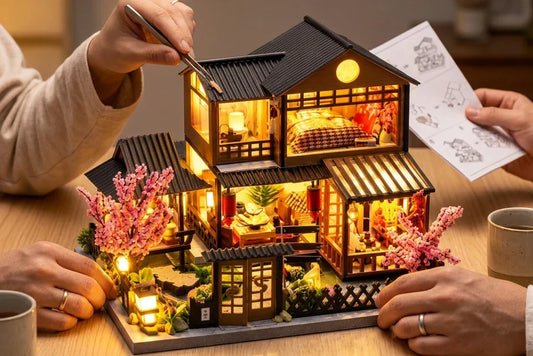 Un couple monte ensemble la maison miniature La Maison aux Cerisiers, allumée sur une table en bois : quatre mains avec alliances, une pince de précision, la notice et des planches de pièces vidées