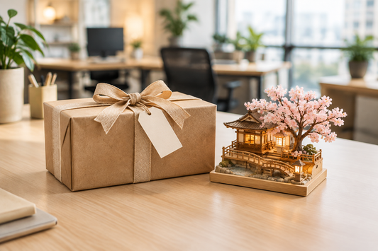 Coffret cadeau en carton kraft avec ruban en tissu et carte vierge, posé sur un bureau lumineux à côté d’une miniature japonaise en bois avec un cerisier en fleurs.