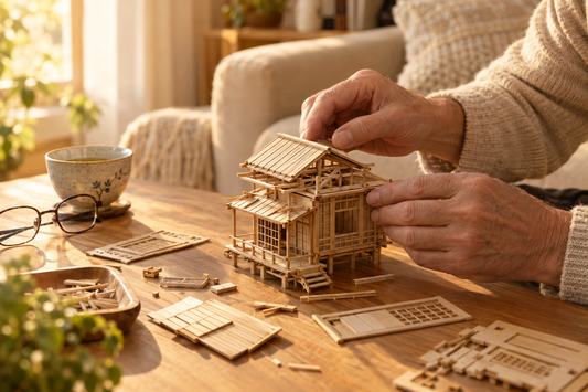 Mains d’une personne d’une soixantaine d’années assemblant une maison miniature japonaise en bois, avec des pièces prédécoupées, une tasse de thé et des lunettes sur une table éclairée par la lumière douce de fin d’après-midi.