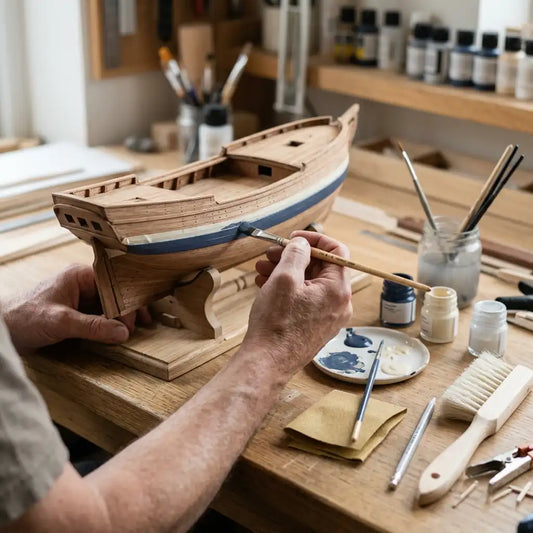 Mains appliquant une peinture bleu marine au pinceau sur la coque d’une maquette de bateau en bois.